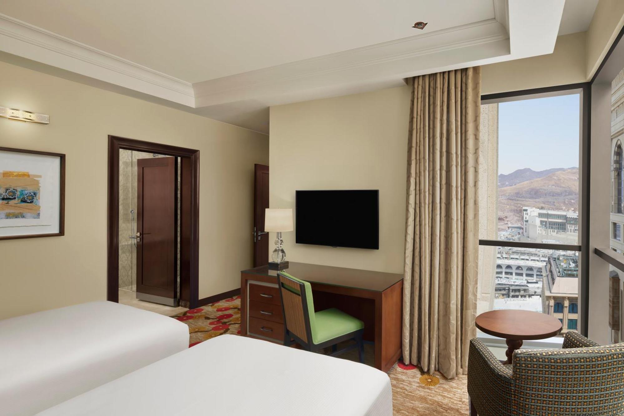 ホテル ヒルトン スイーツ マッカ メッカ エクステリア 写真 The photo shows a spacious hotel room featuring two beds, a desk with a green chair, and a flat-screen TV mounted on the wall. There's a large window with curtains that lets in natural light and offers a view of the outside. The decor is modern and w